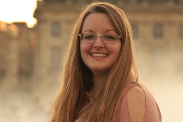 A portrait of a smiling Rose Ling, a white woman with long hair and glasses, stood side on in front of old yellow brick buildings